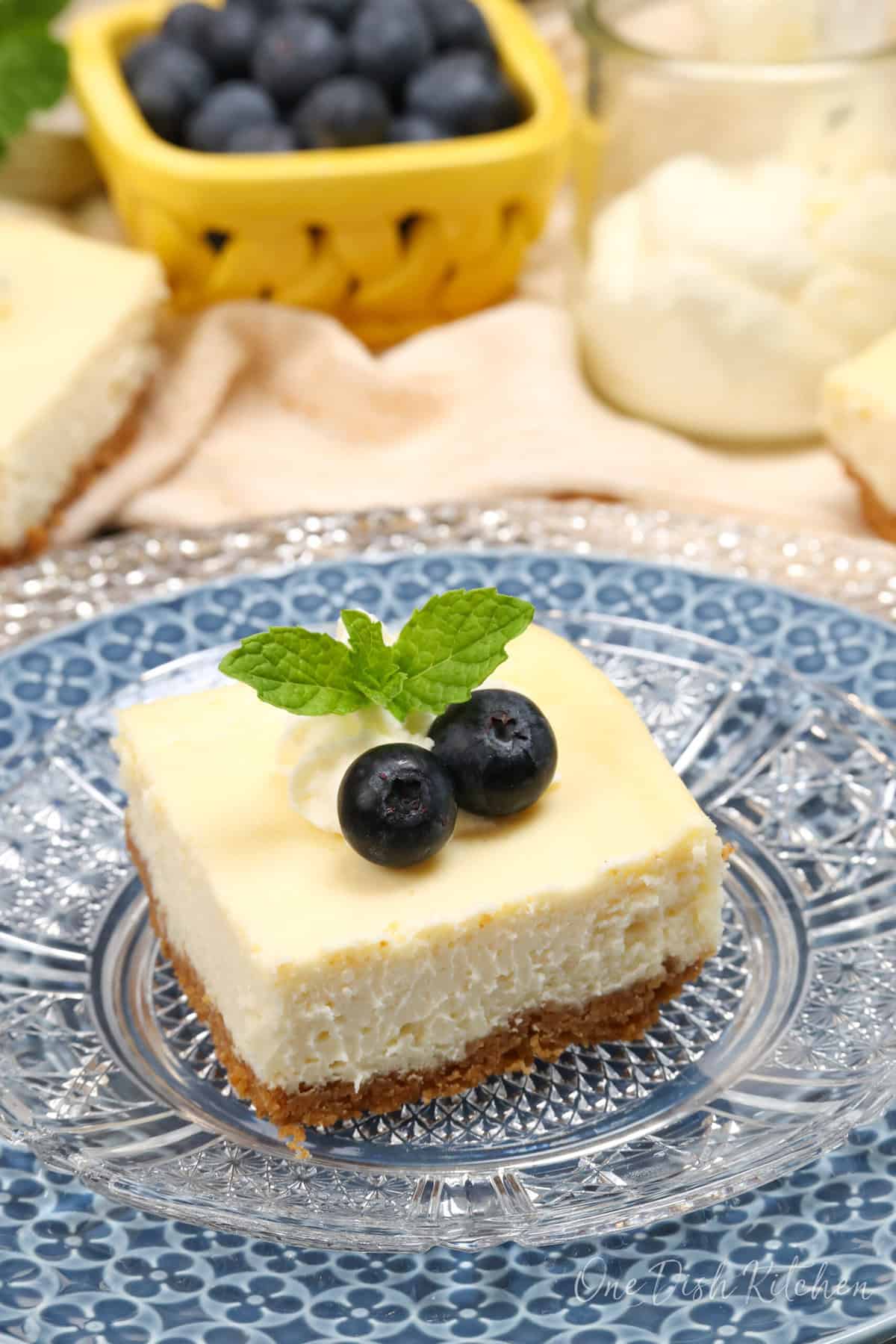 a cheesecake square topped with blueberries and fresh mint next to three other cheesecake bars.