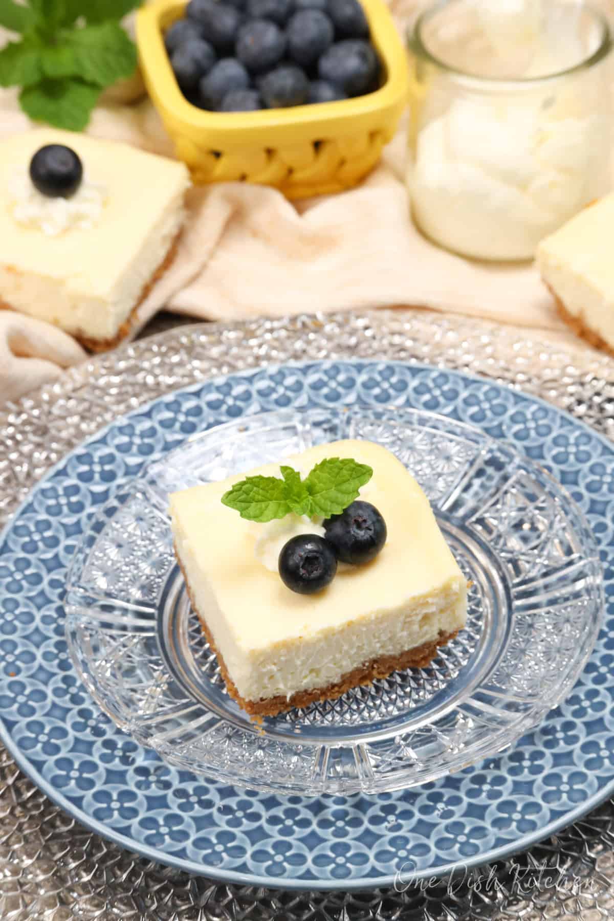 one cheesecake bar on a blue plate next to another bar, a bowl of blueberries, and whipped cream.