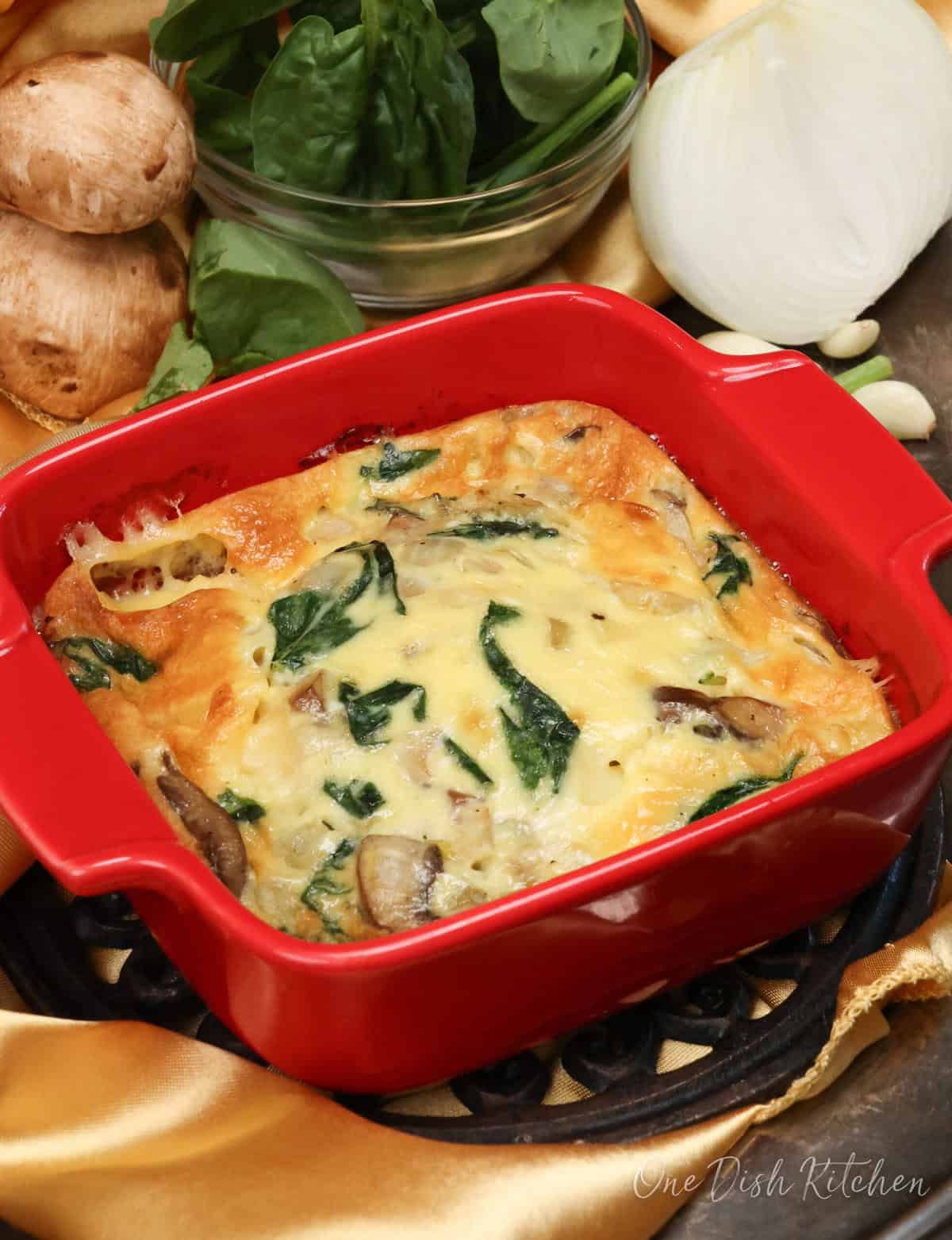 a crustless quiche with spinach and mushrooms in a red square baking dish.