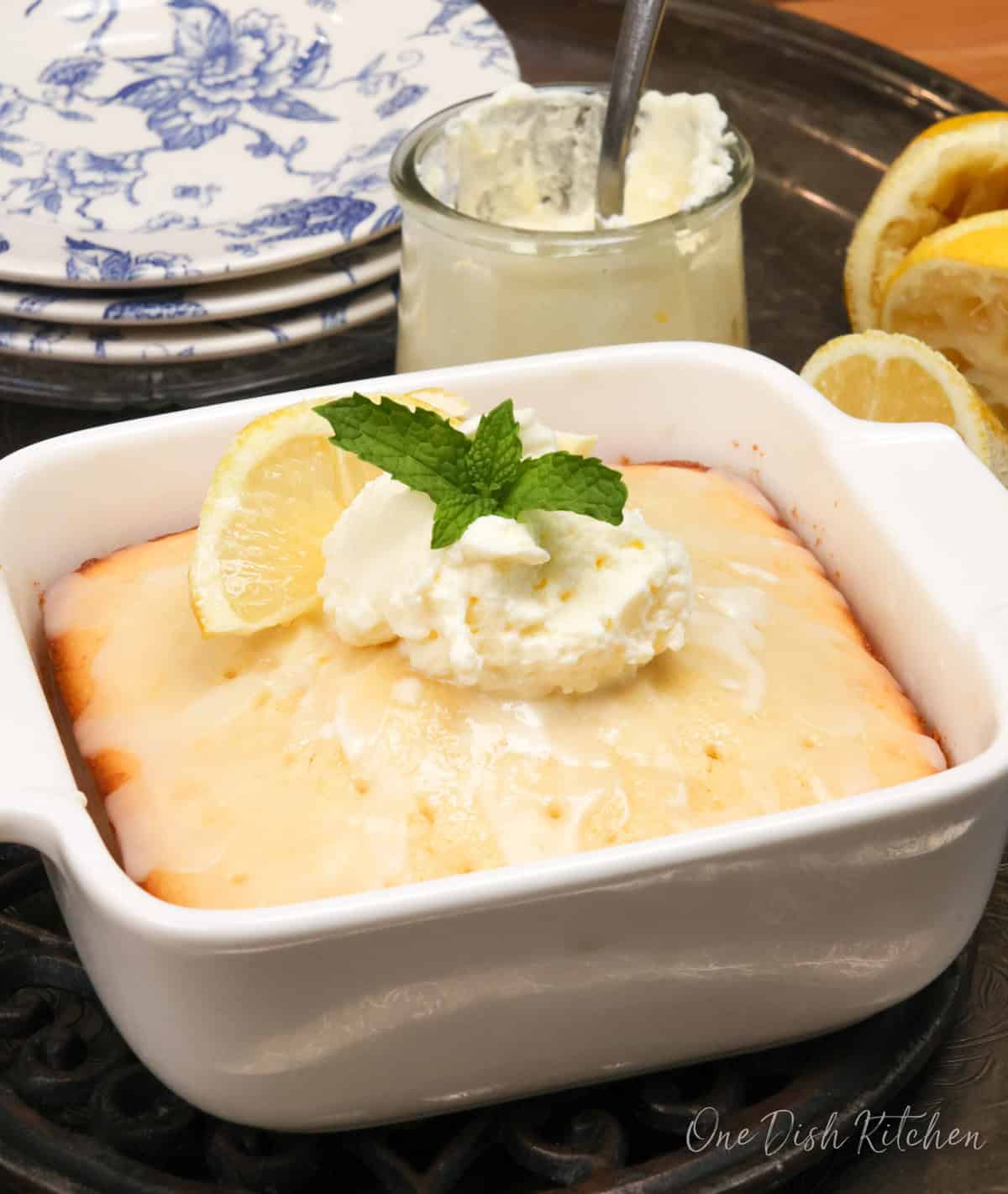 a small lemon cake in a square baking dish next to a jar of whipped cream and three plates.