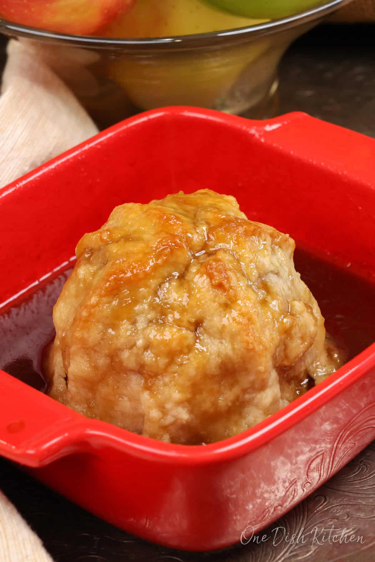 an apple dumpling in a red baking dish with sauce surrounding the apple.