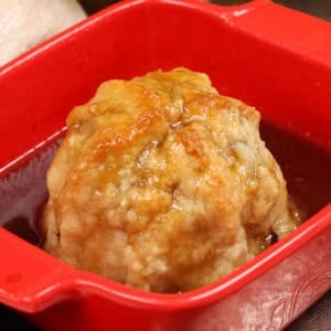 an apple dumpling in a red baking dish with sauce surrounding the apple.