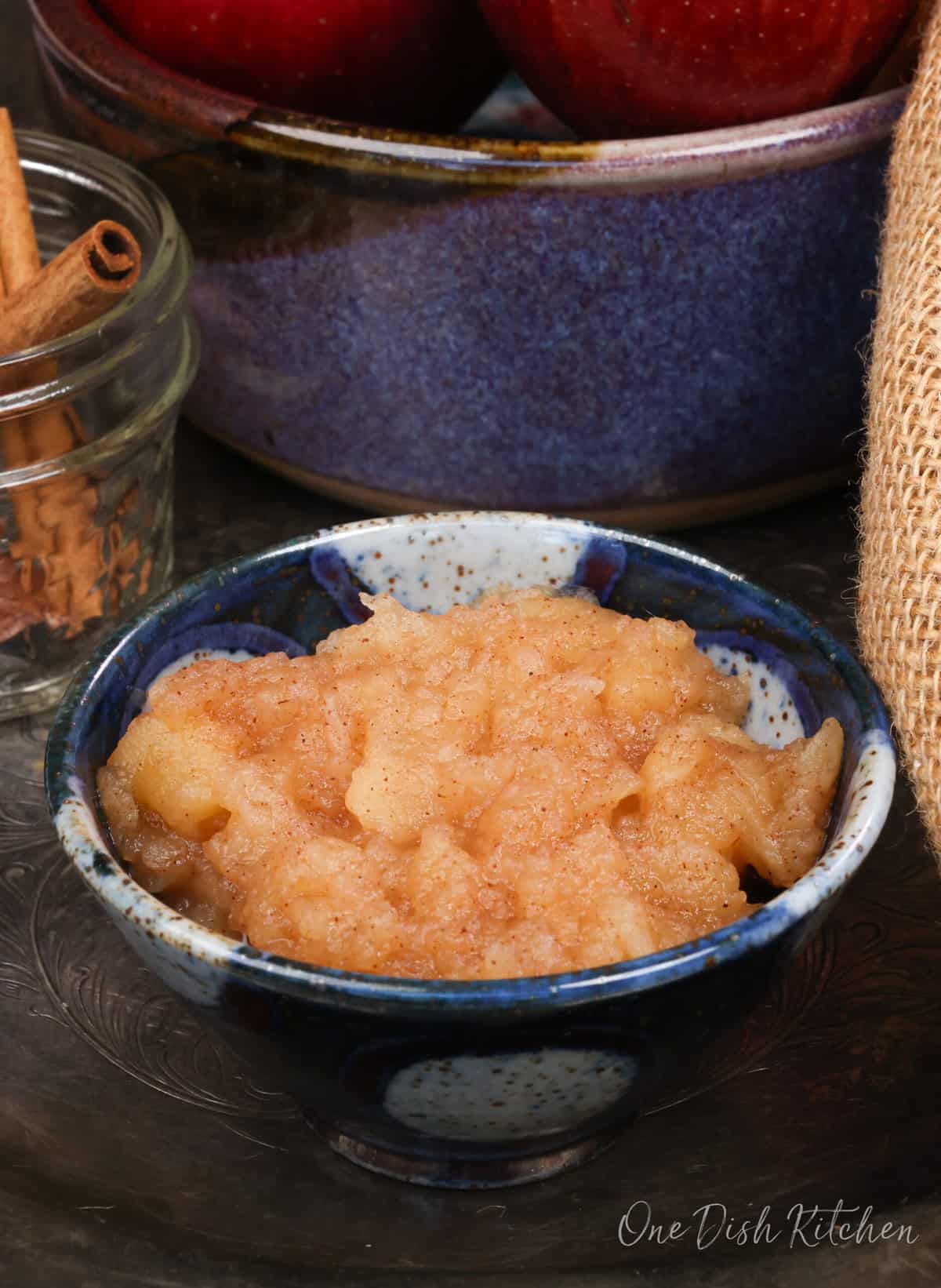 a small bowl of applesauce on a silver tray next to a jar of cinnamon sticks and fresh apples.