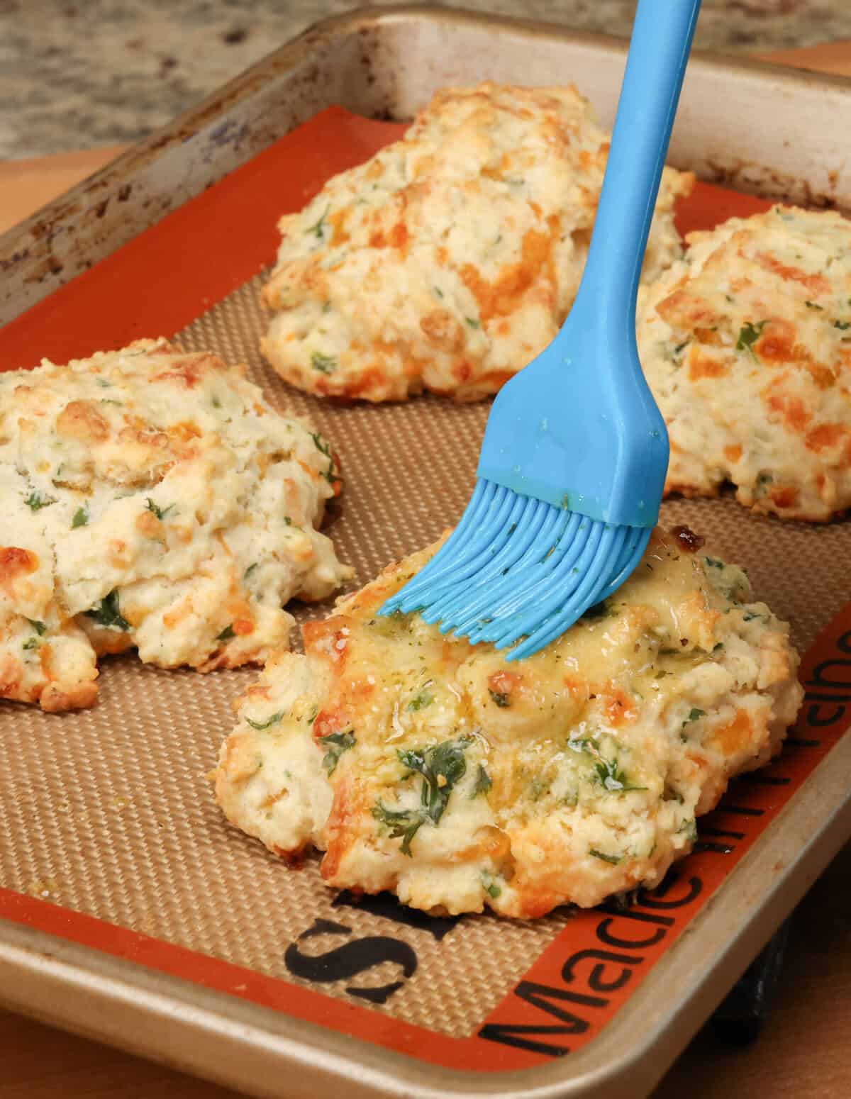 brushing garlic butter over the tops of cheddar biscuits on a baking sheet.