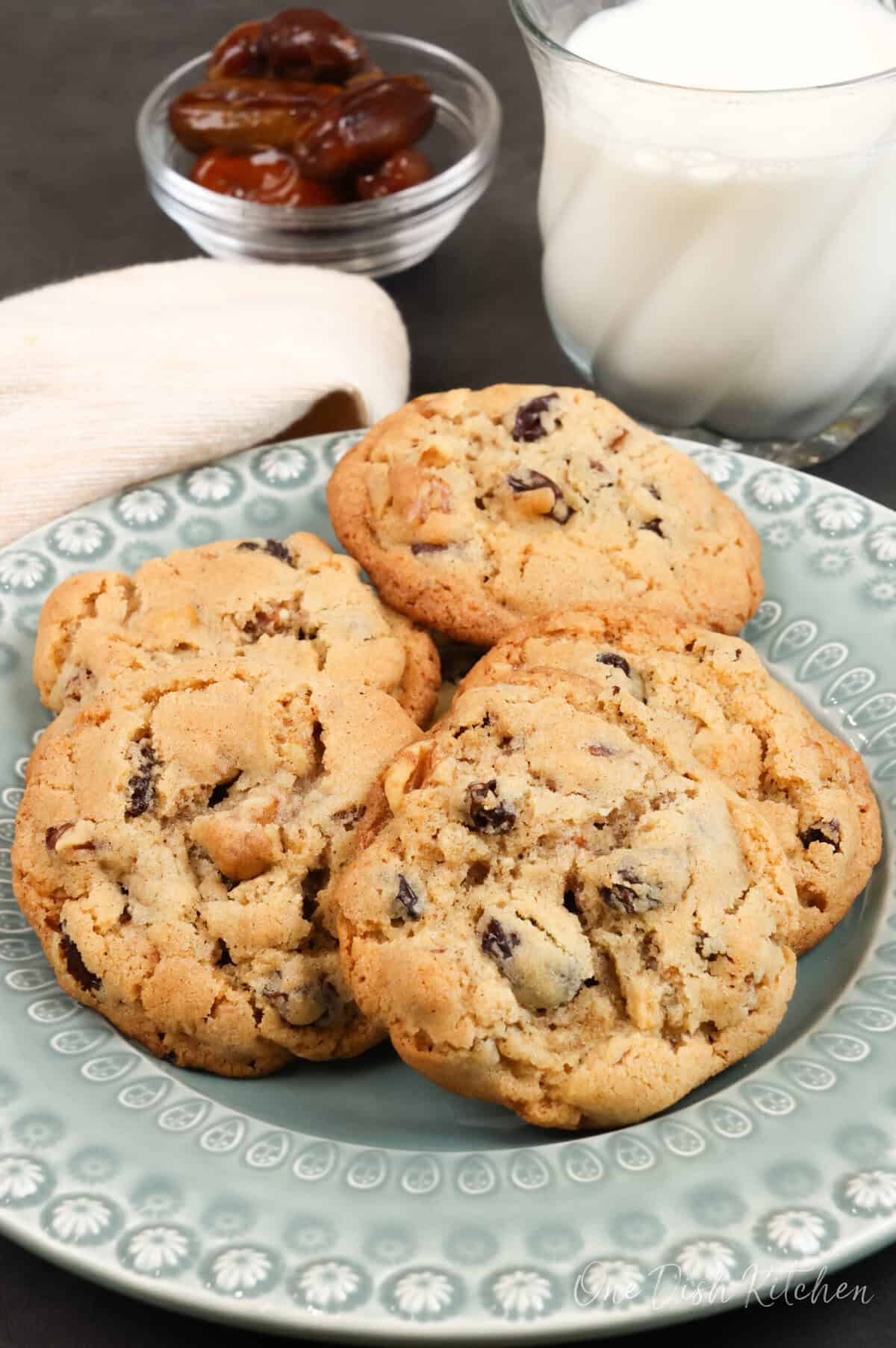 six hermit cookies on a green plate next to a bowl of dates and a glass of milk.