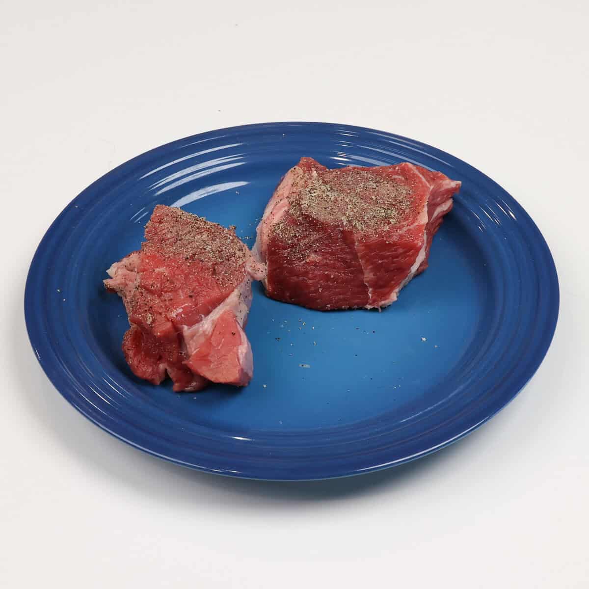 two pieces of beef seasoned with salt and pepper on a blue plate.