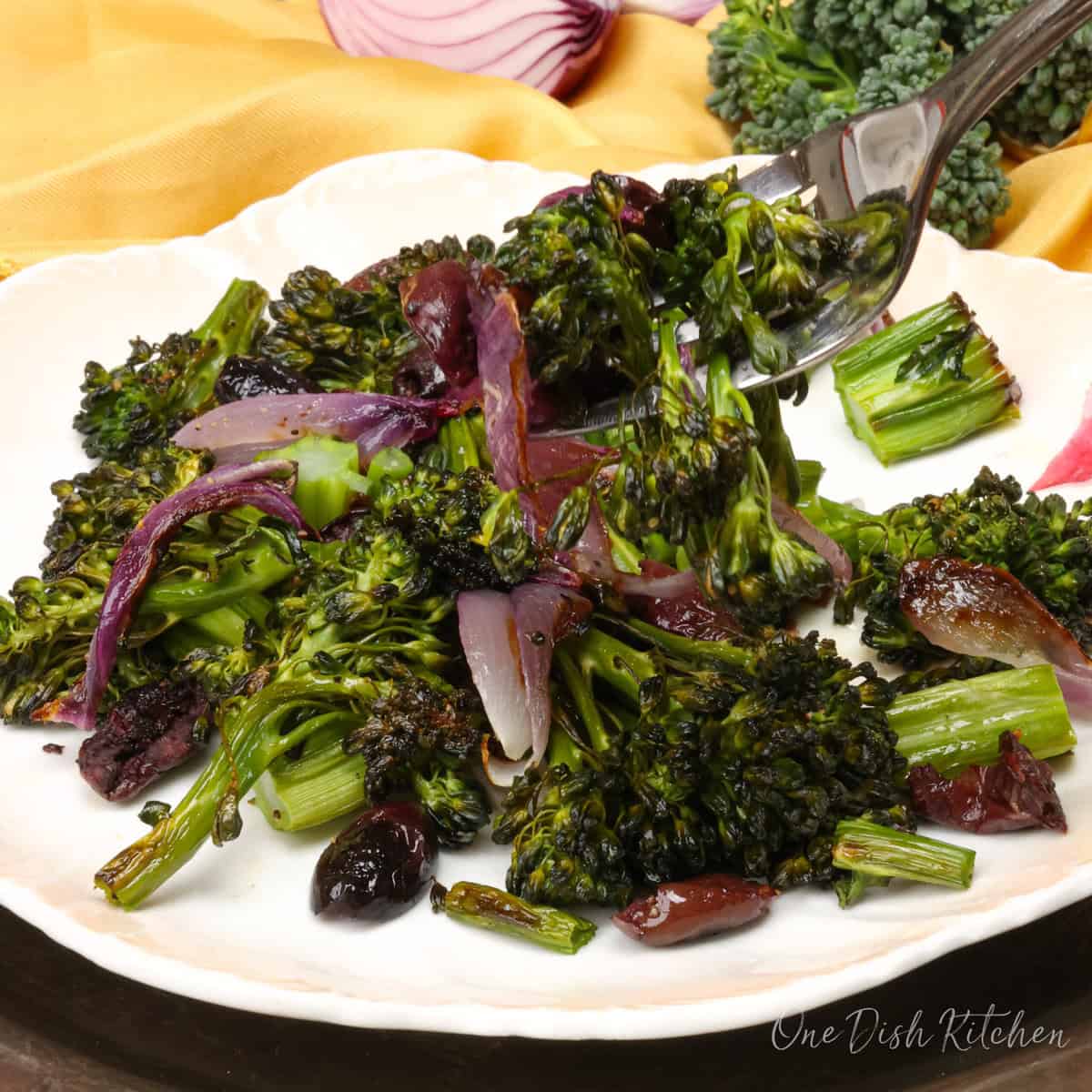 roasted broccolini on a fork above a plate full of more broccolini, onions, and olives.