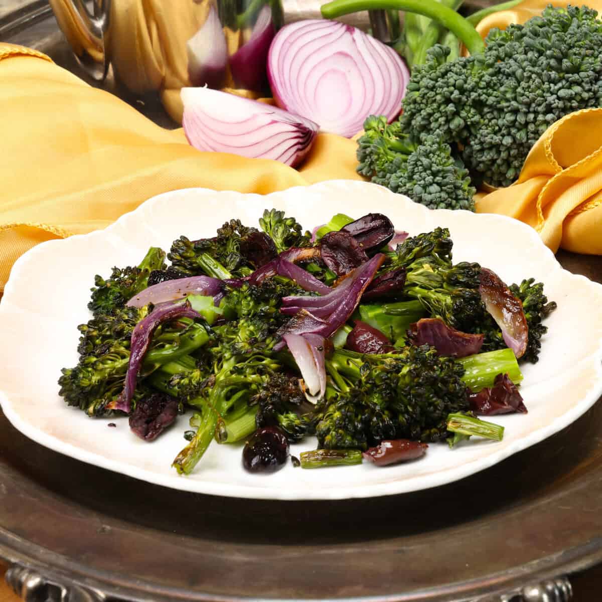 a plate of roasted broccolini with red onions on a silver tray next to a gold napkin.