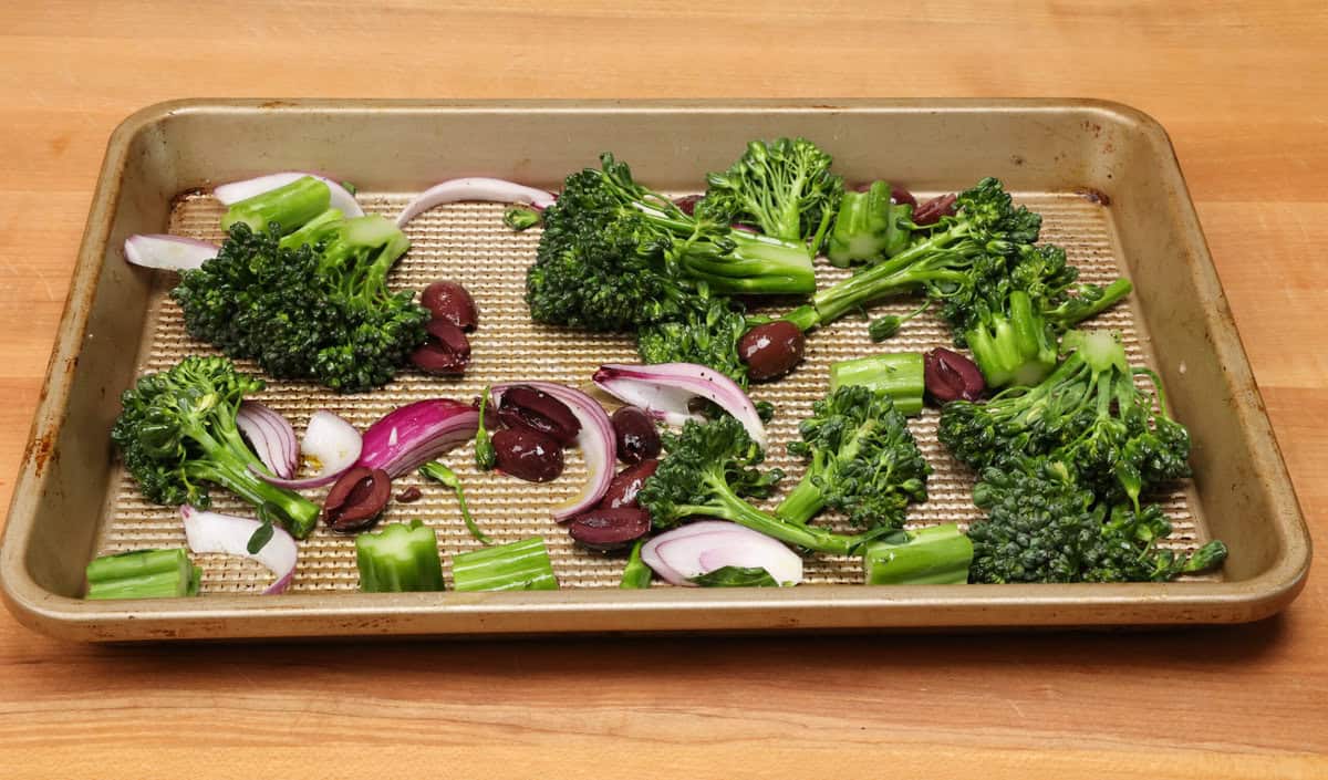 uncooked broccolini, olives and onions on a baking sheet.