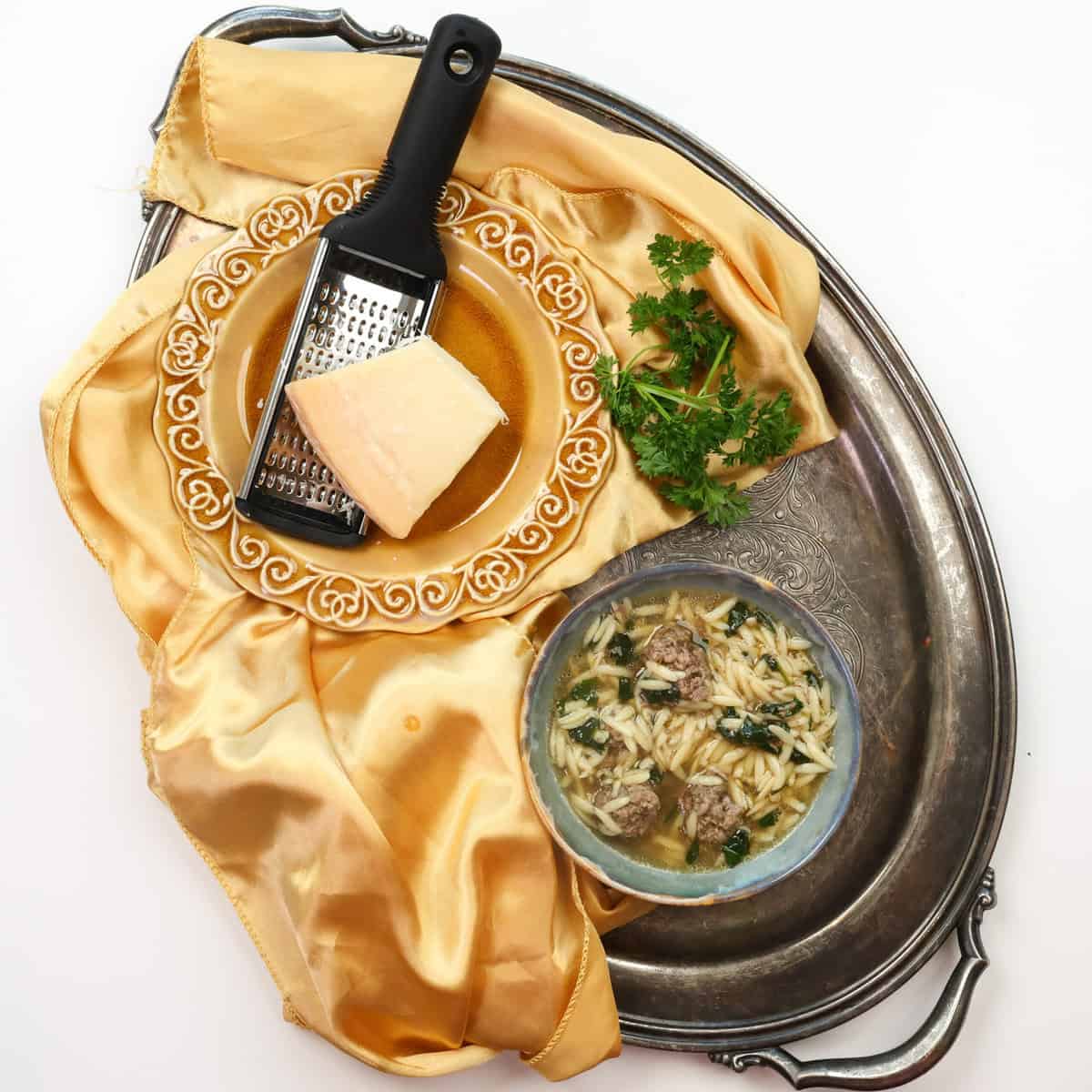 a bowl of Italian Wedding Soup on a silver tray next to a gold napkin and parmesan cheese.