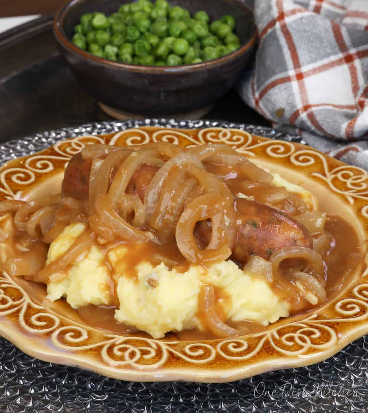 bangers and mash on a gold plate next to a bowl of peas.