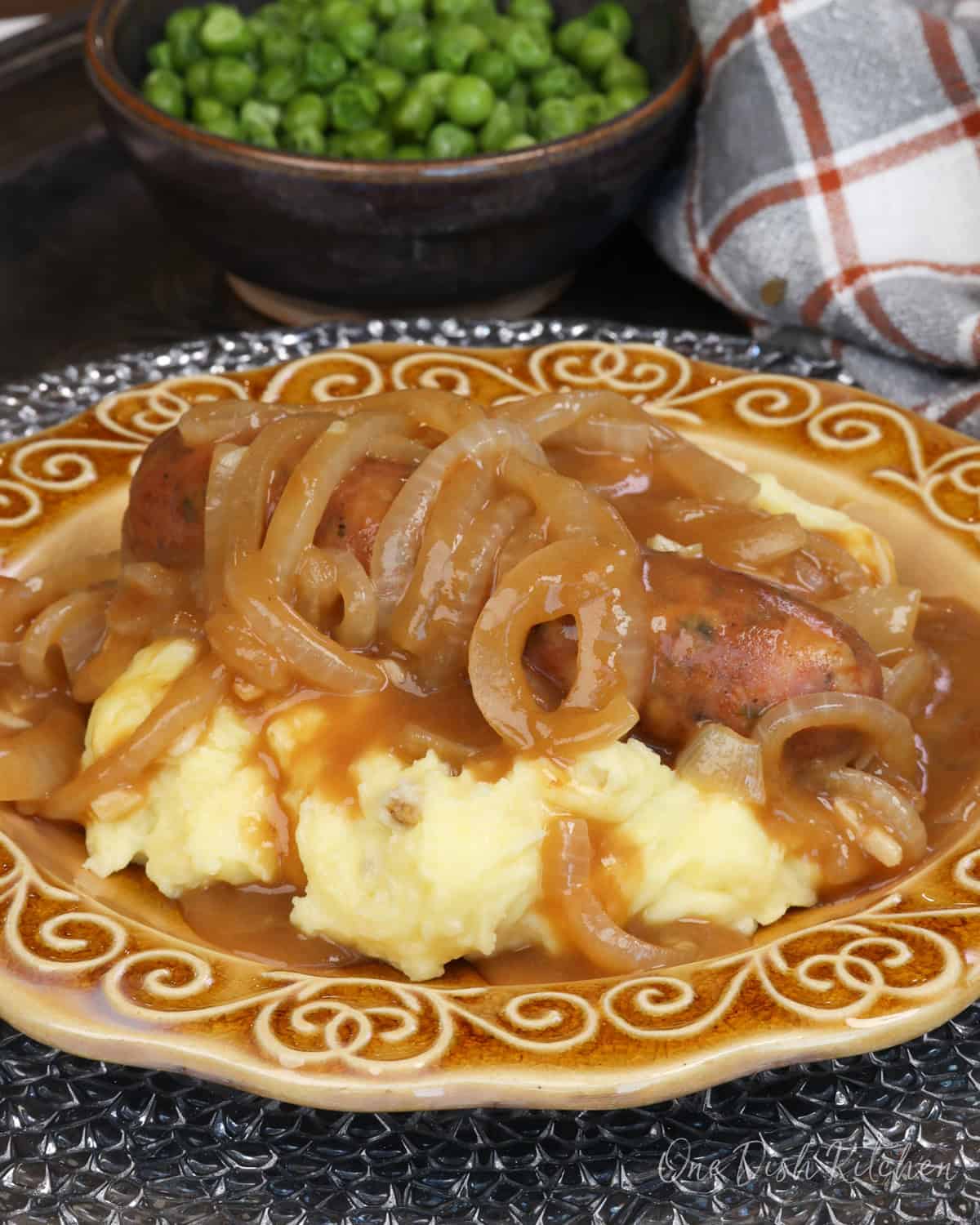 bangers and mash on a gold plate next to a bowl of peas.