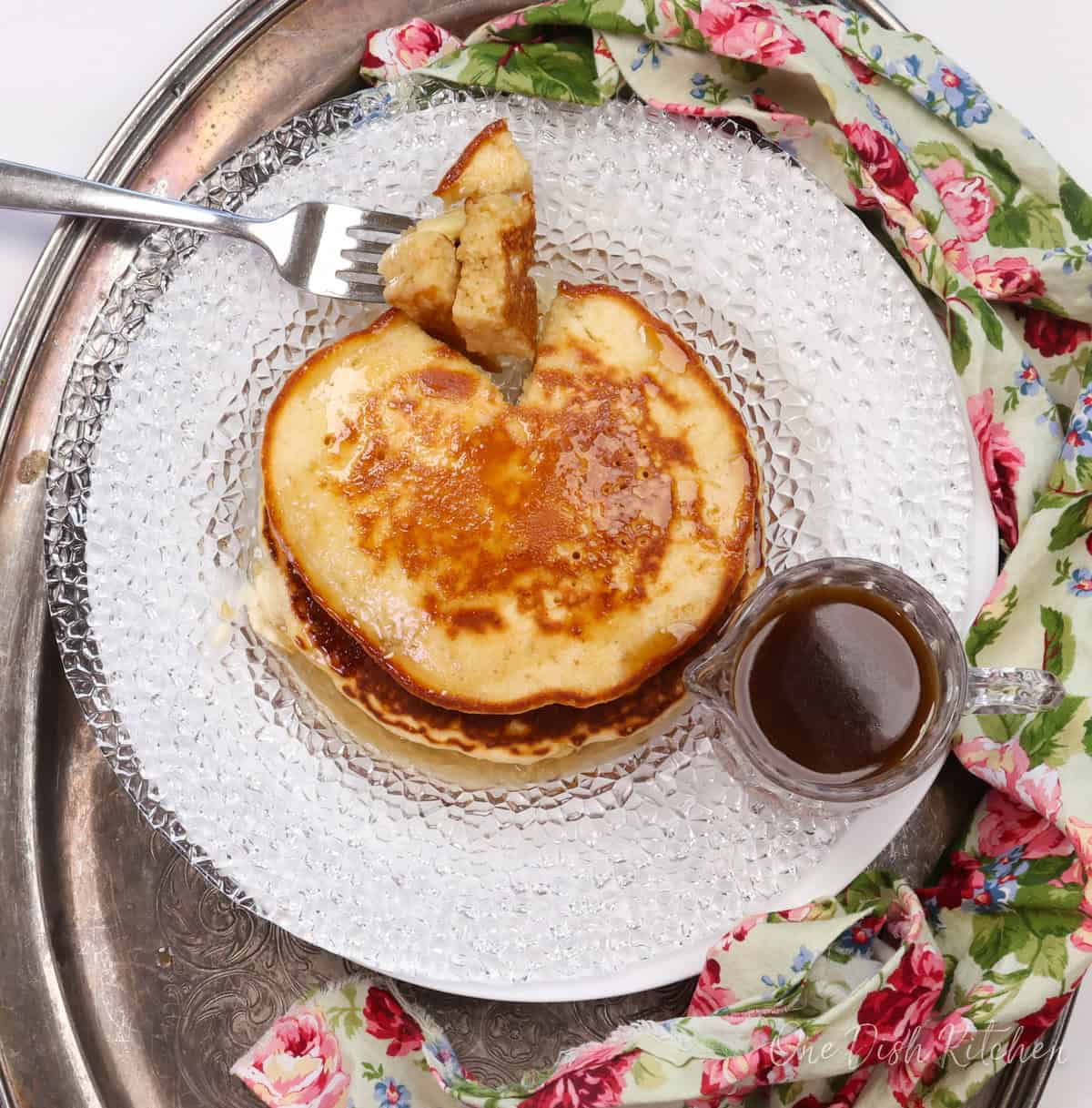 pancakes on a plate next to a pink floral napkin and a jar of syrup.