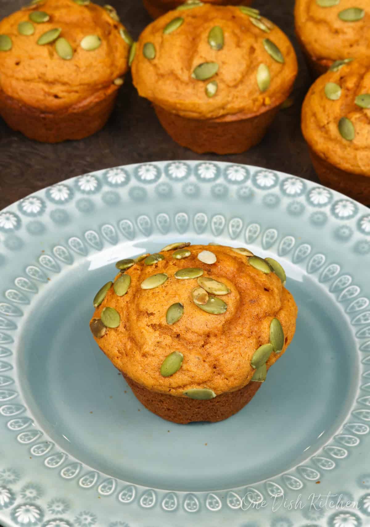 one pumpkin muffin on a green plate next to 5 muffins on a tray.