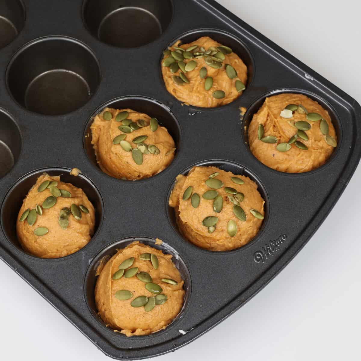 unbaked pumpkin spice muffins topped with pumpkin seeds in a muffin tin.