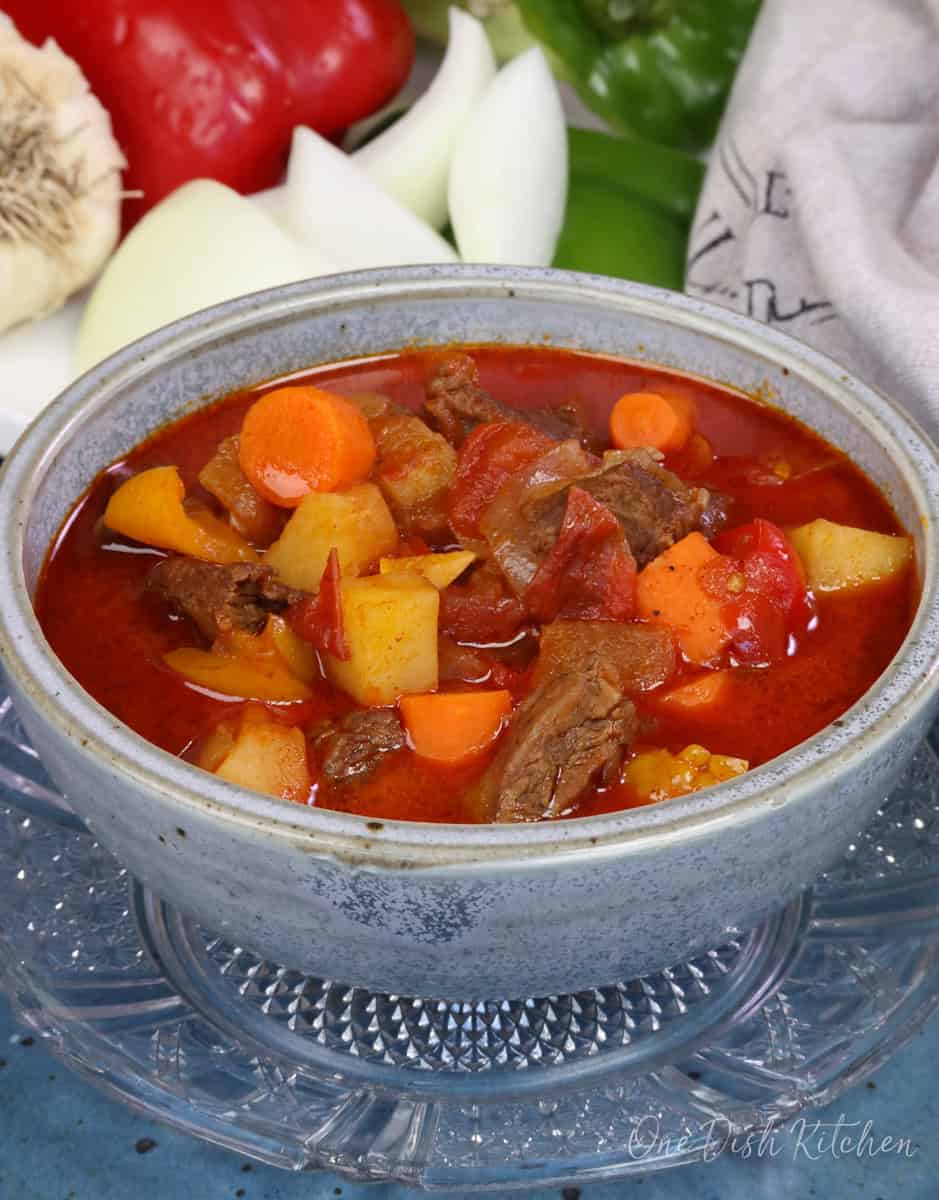 a blue bowl filled with hungarian goulash next to fresh vegetables.