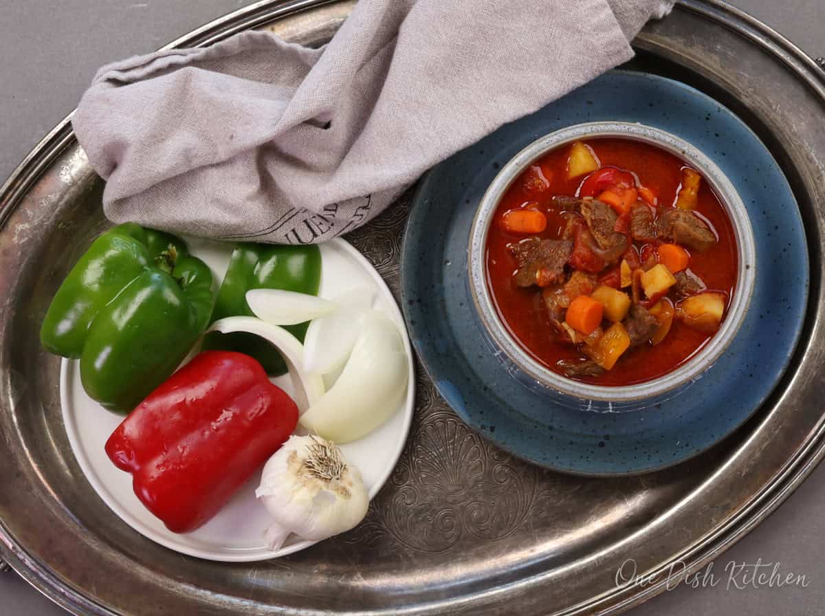 a bowl of hungarian goulash on a silver tray next to a brown napkin and a plate of vegetables.