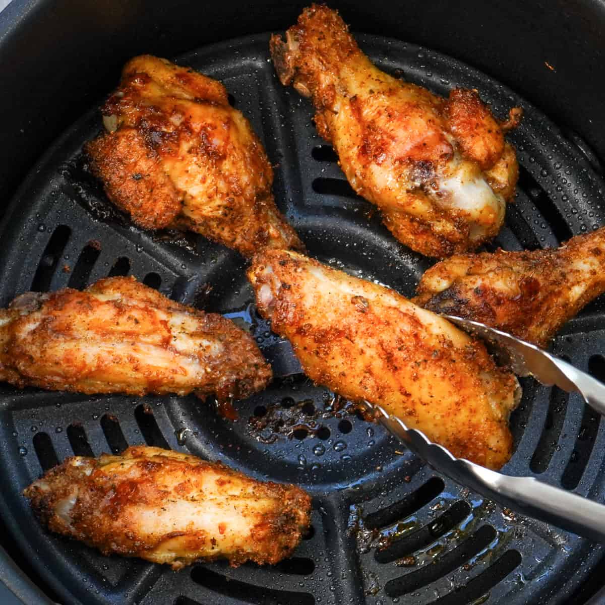 tongs lifting a chicken wing out of an air fryer.