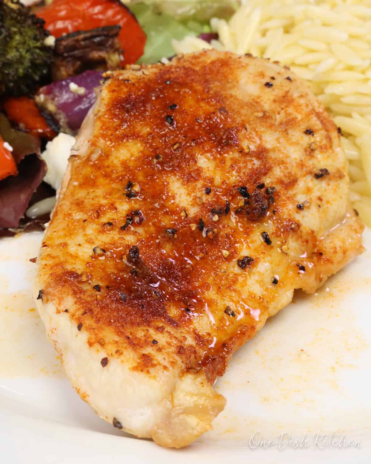 a baked chicken breast on a white plate next to salad and cooked orzo.