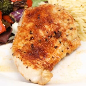 a baked chicken breast on a white plate next to salad and cooked orzo.