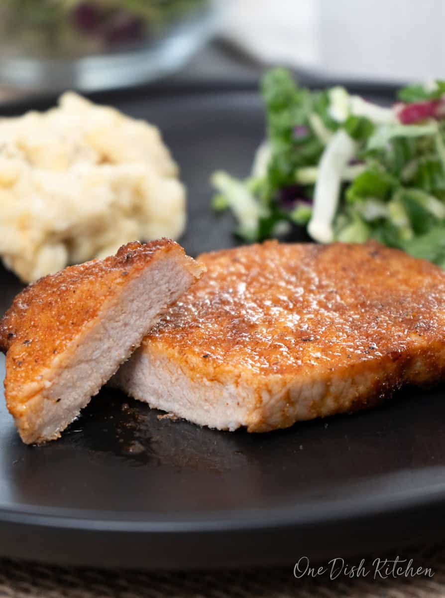 a pork chop on a black plate, sliced and served with mashed potatoes and a salad.