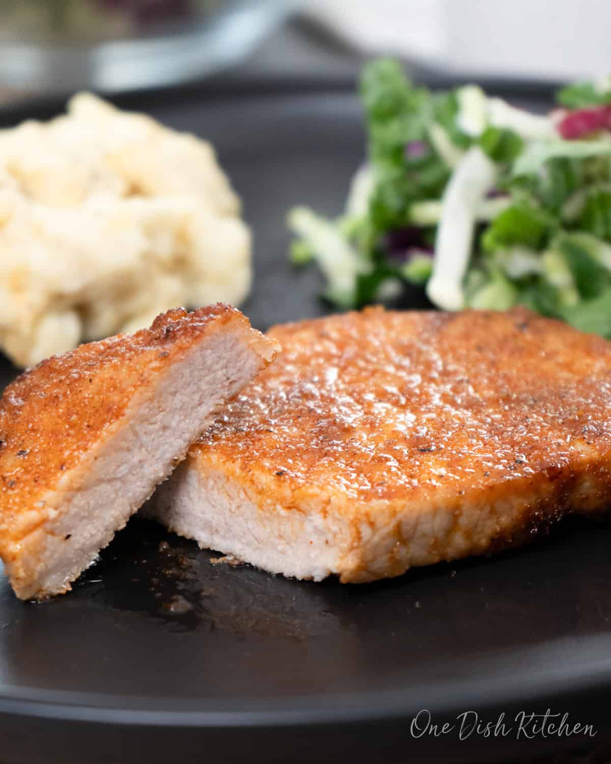 a pork chop on a black plate, sliced and served with mashed potatoes and a salad.