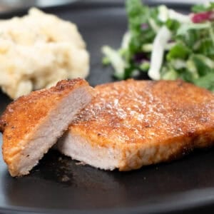 a pork chop on a black plate, sliced and served with mashed potatoes and a salad.