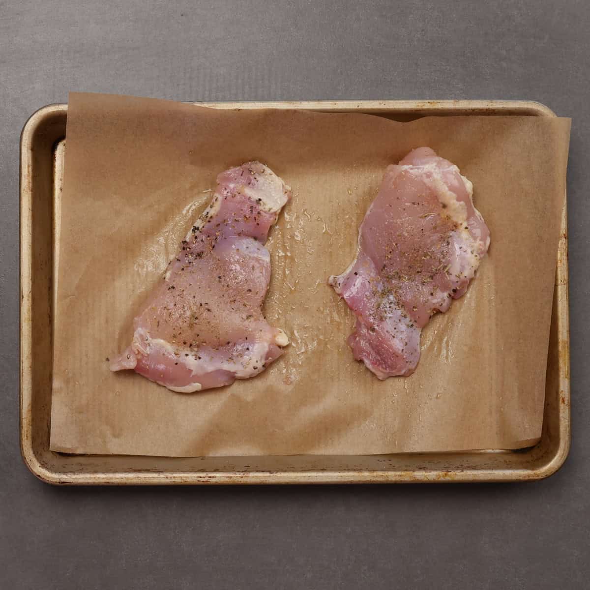 two uncooked boneless skinless chicken thighs on a baking sheet.