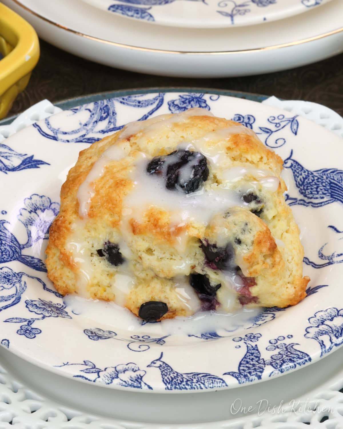 two blueberry scones on plates next to a yellow bowl of fresh blueberries.