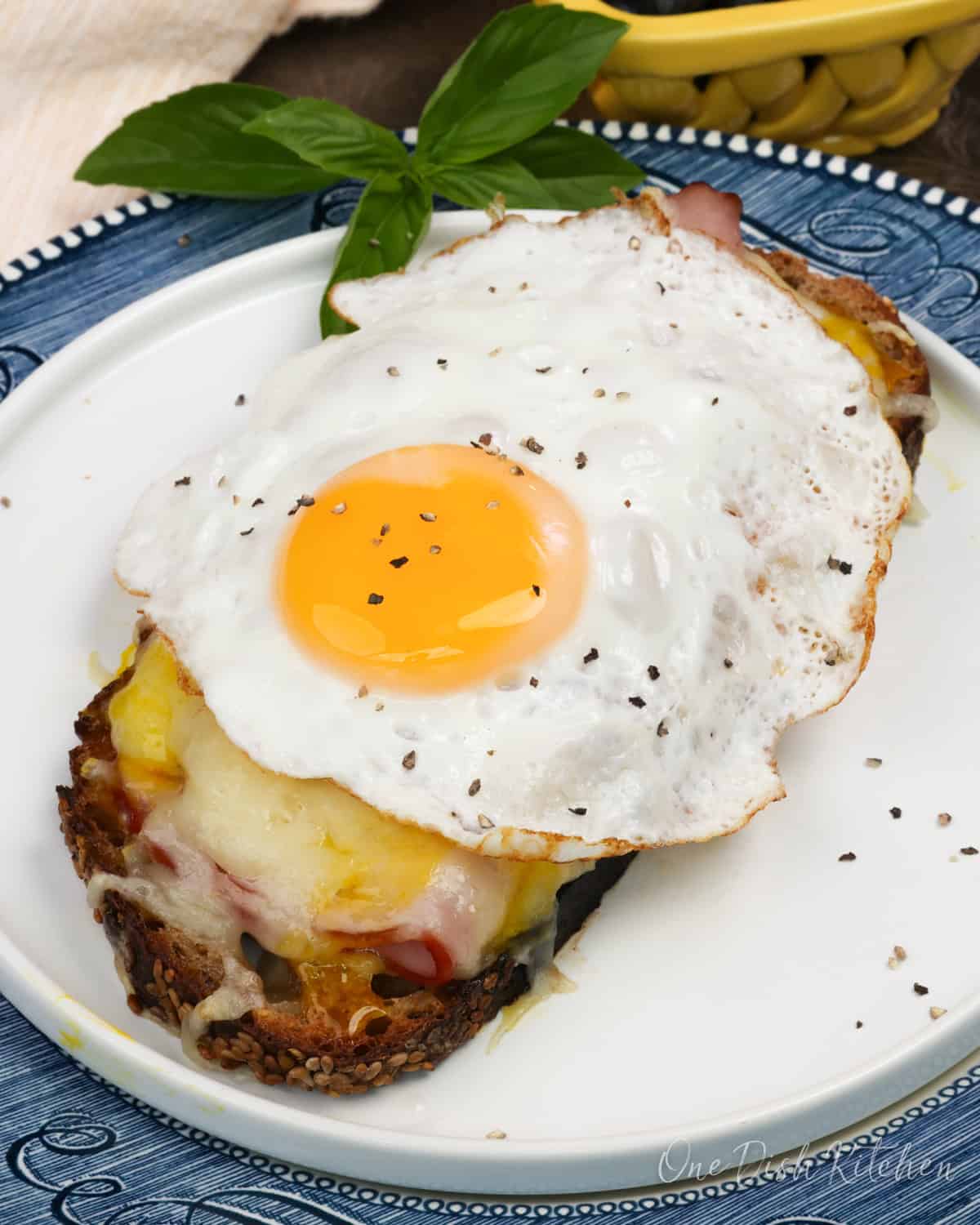 an open faced croque madame on a white plate next to fresh basil.
