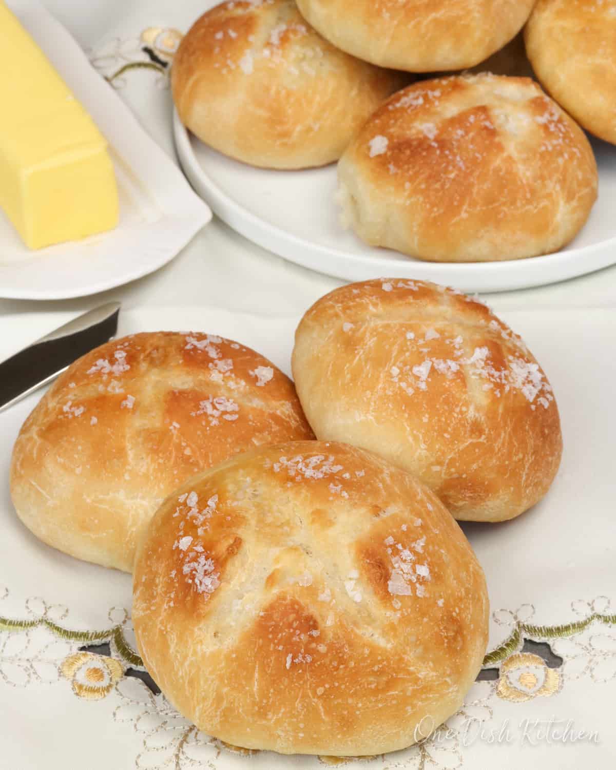 a small batch of french bread dinner rolls on a tablecloth next to a stick of butter.
