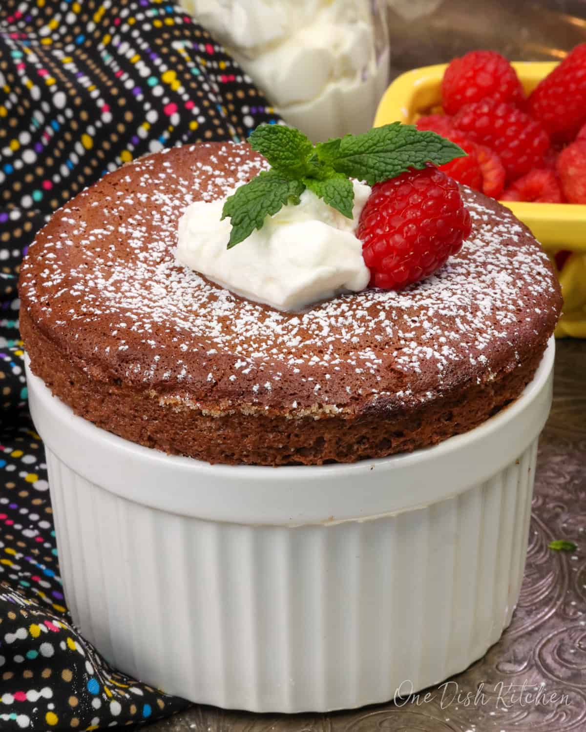 chocolate soufflé for one baked in a white ramekin topped with whipped cream and a raspberry.