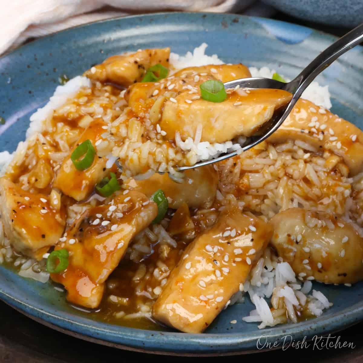 a single serving of orange chicken on a plate over white rice and topped with sesame seeds and chopped green onions.