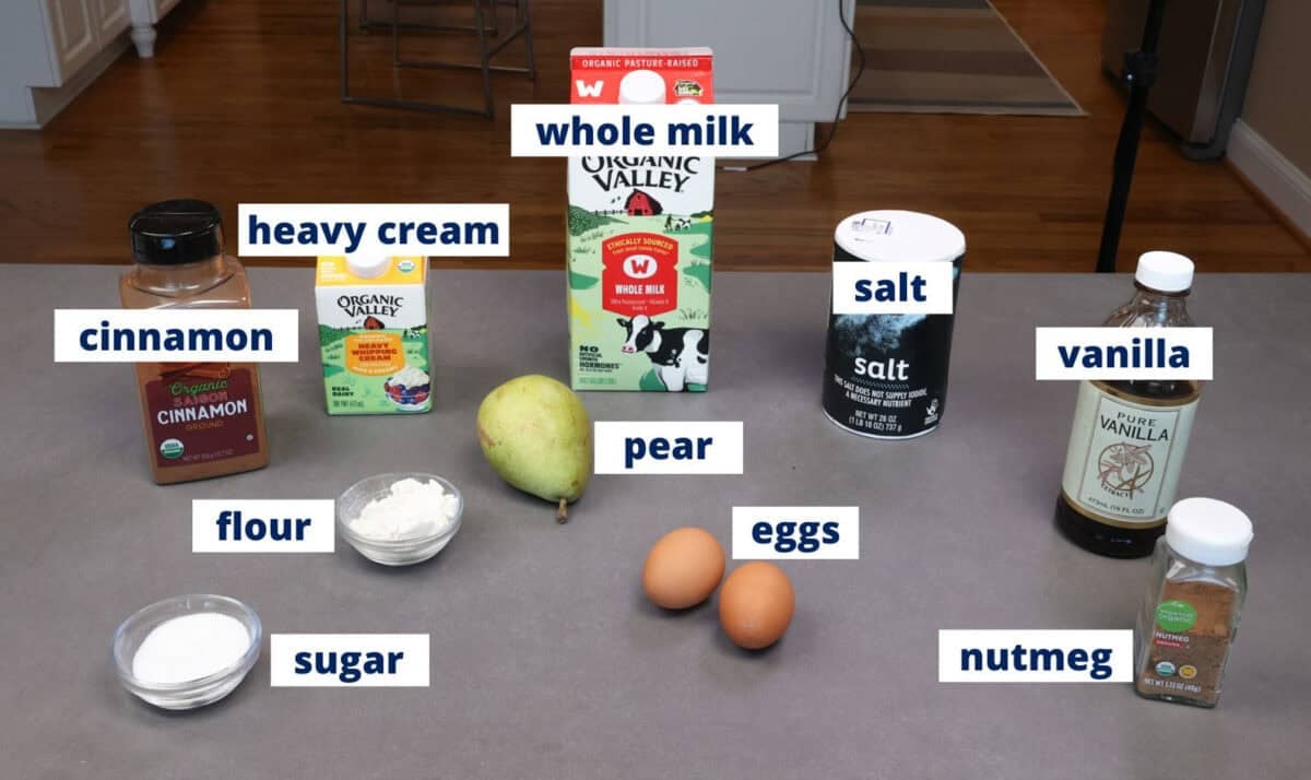 ingredients in a pear clafoutis including eggs, a pear, milk, cream, sugar, and flour arranged on a kitchen counter.