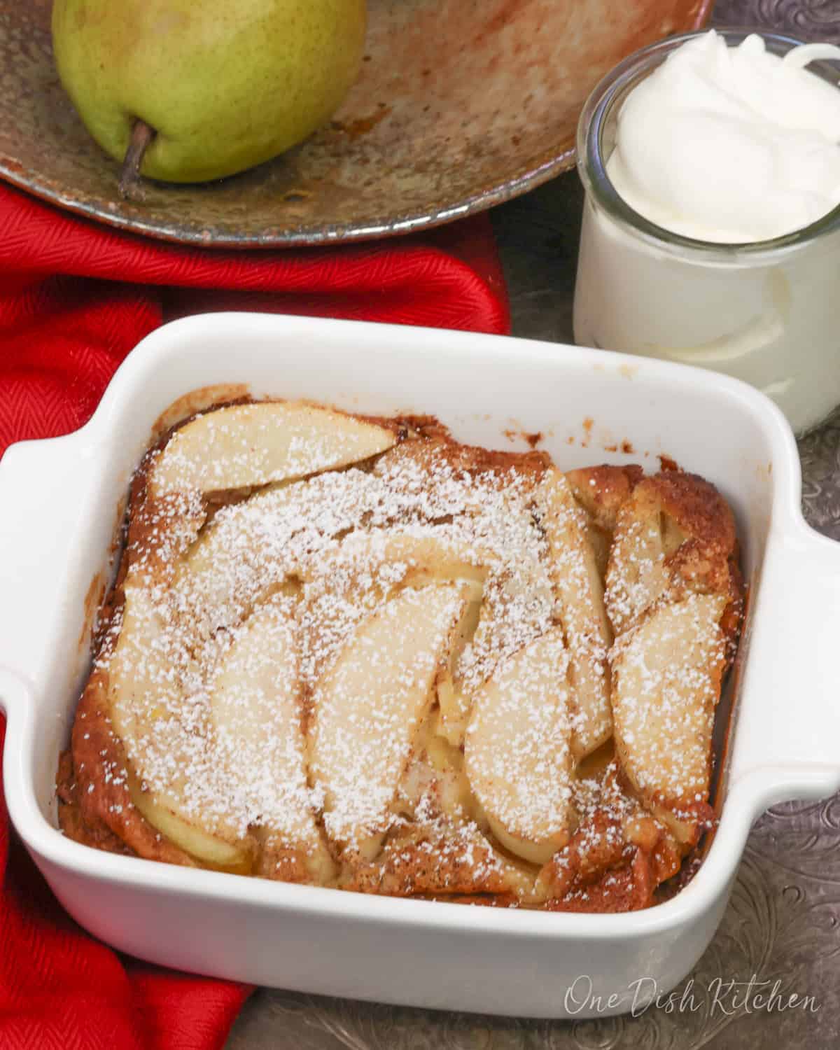 a single serving pear clafoutis in a small baking dish next to a bowl of pears.