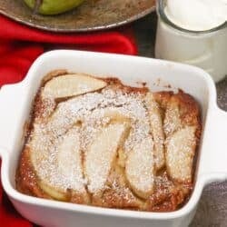 a single serving pear clafoutis in a small baking dish next to a bowl of pears.